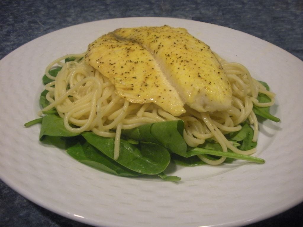 A white plate on a blue counter top with spinach, spaghetti, and tilapia.