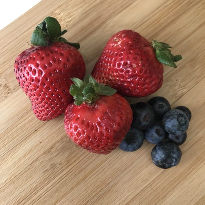 3 strawberries and a bunch of blueberries on a light wooden cutting board.